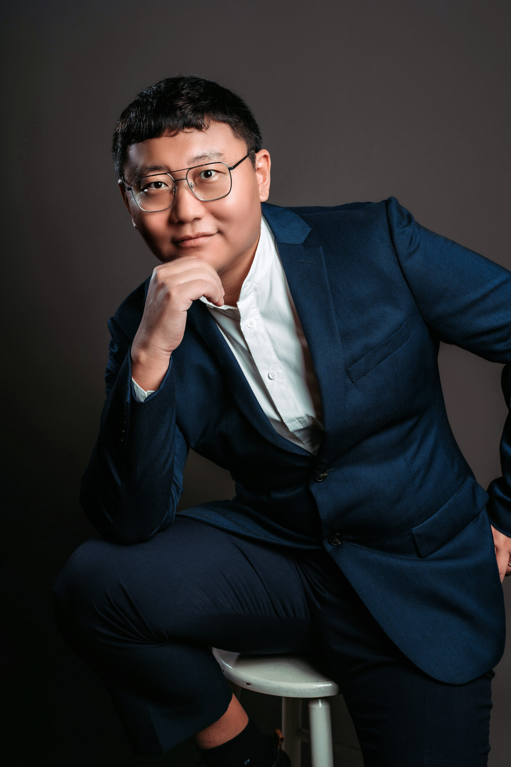 Man in navy suit sitting on stool, hand on chin